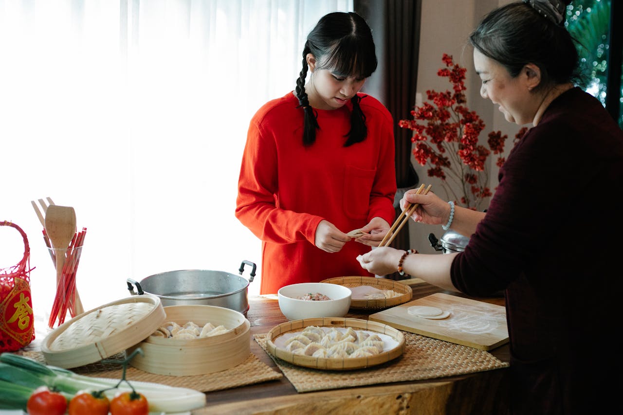 Cheerful ethnic grandma with attentive teen preparing dumplings at table with traditional steamers during New Year holiday at home