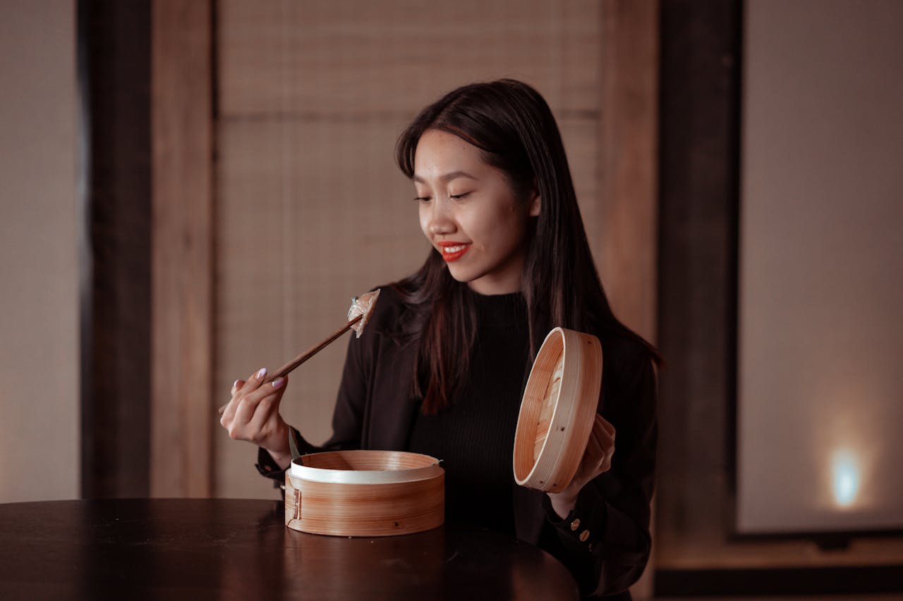 Young woman enjoying dumplings with chopsticks from a bamboo steamer indoors.